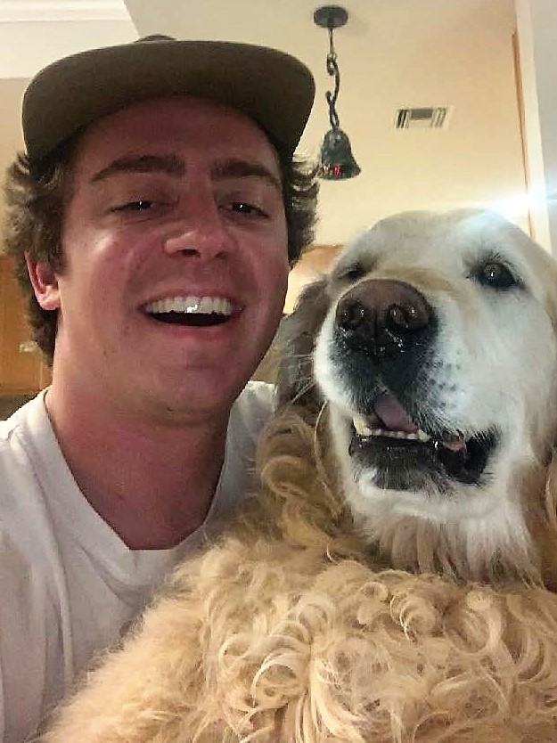 Smiling man with curly hair wearing a cap and  taking a selfie with his dog