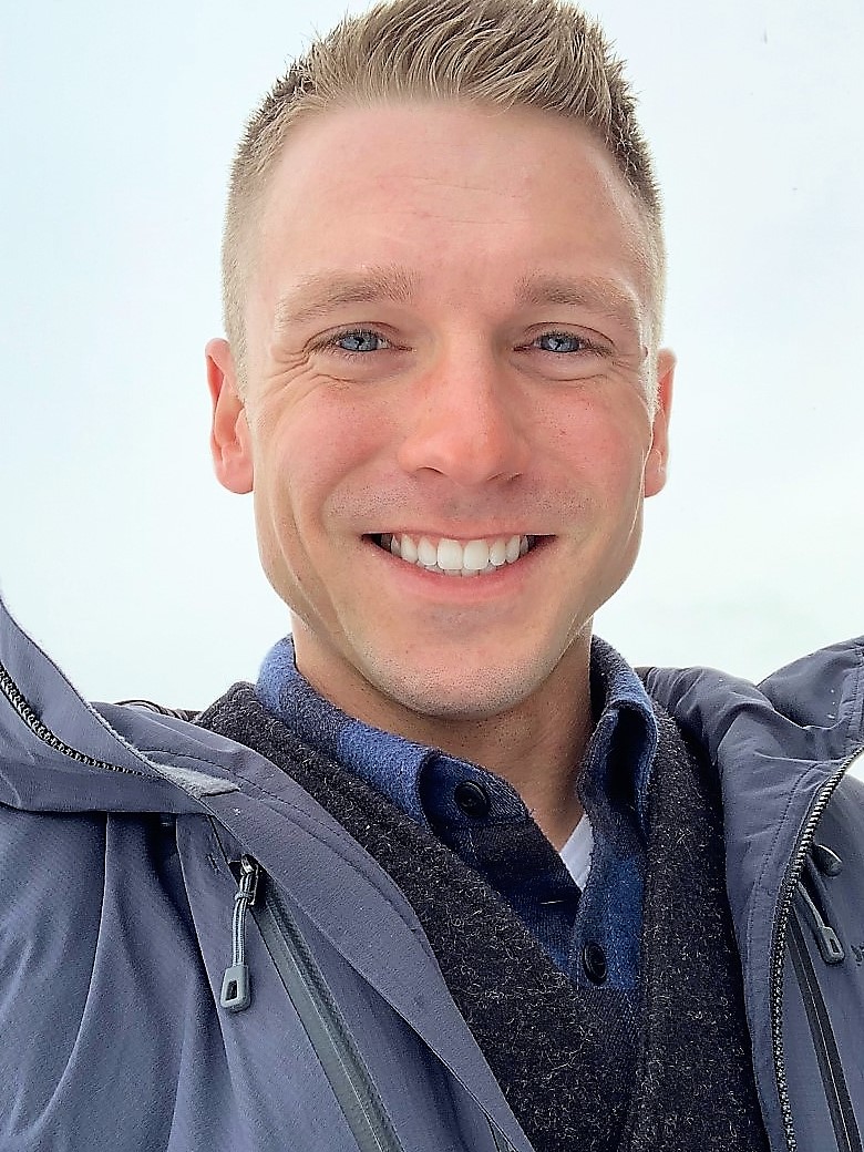 A man with short blonde hair is smiling at the camera while in the outdoors