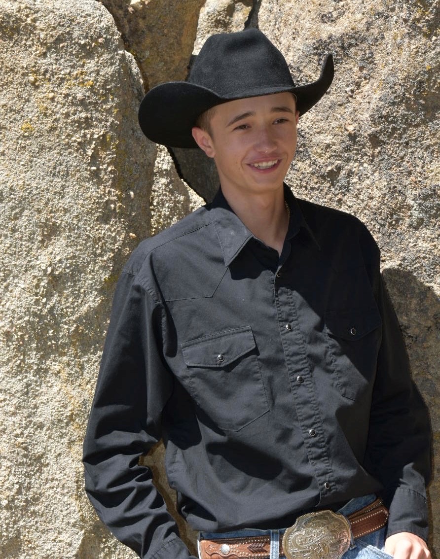 a young man in black western wear leaning against a mountain