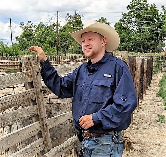 A man in a cowboy hat and blue shirt with one hand on a rustic wooden fence post