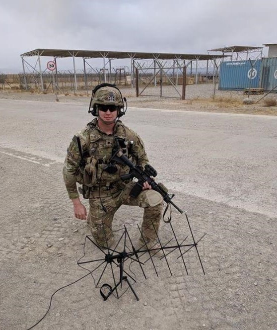 A man in military fatigues is shown kneeling on sandy ground