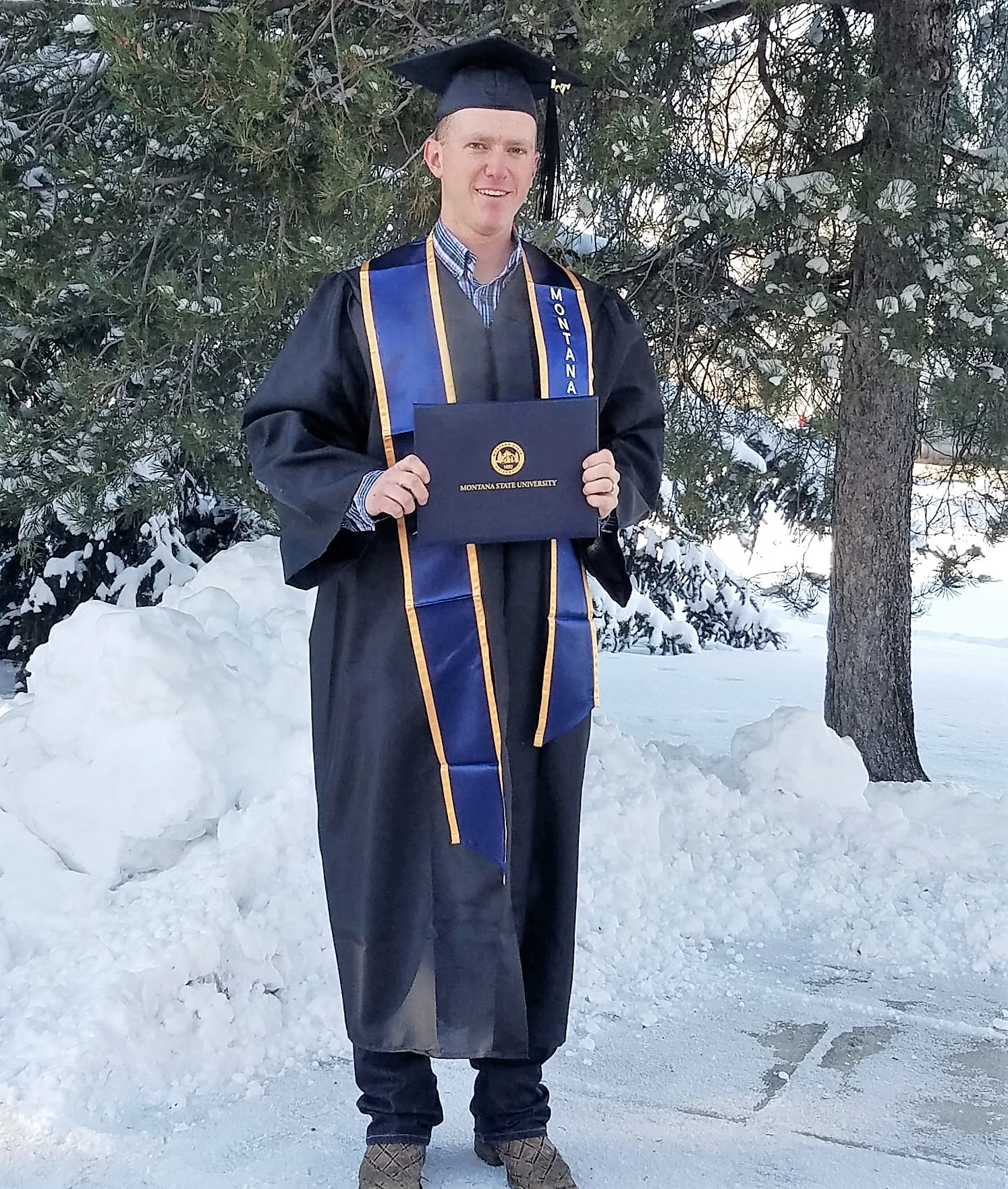 a man wearing montana state commencement regalia holds a diploma