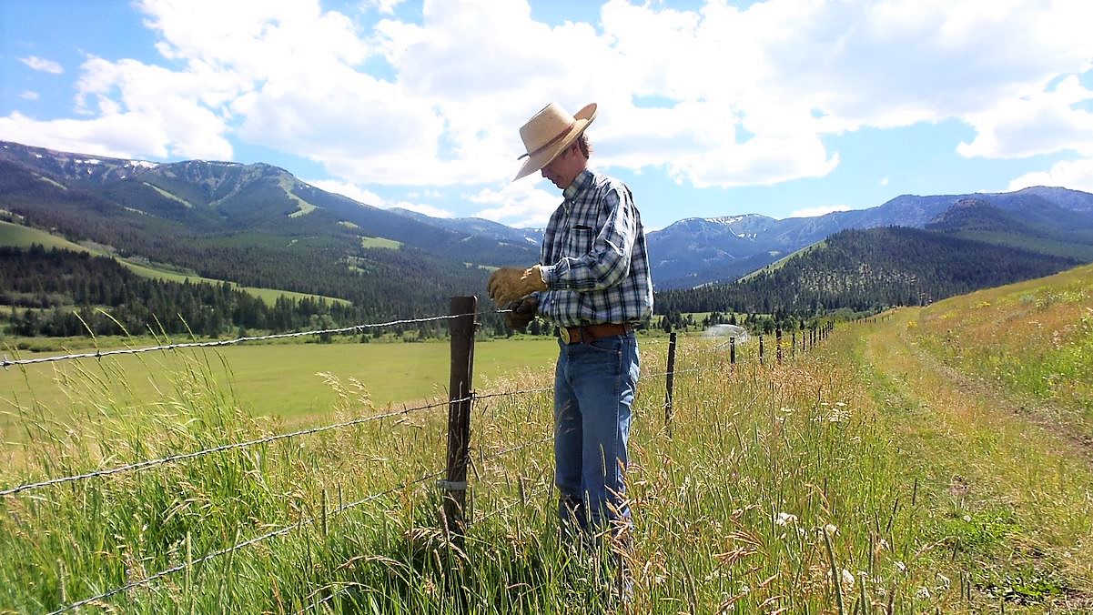 A young man in a cowboy hat and western shirt is shown repairing pasture fence with mountains in the background and blue sky and white clouds overhead.