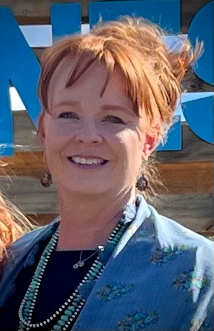 a woman with red hair in an updo wearing a denim shirt with flowers on it smiles at the camera